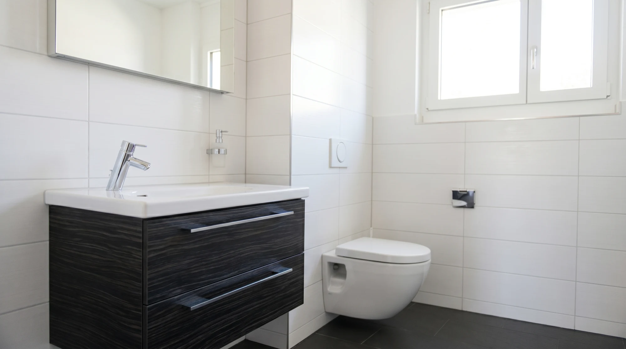 Saniertes Badezimmer von Zemp Sanitär AG Luzern — Waschtisch mit dunkler Holzfront, verchromte Armatur, Wandspiegel und wandhängendes WC, helle Fliesen