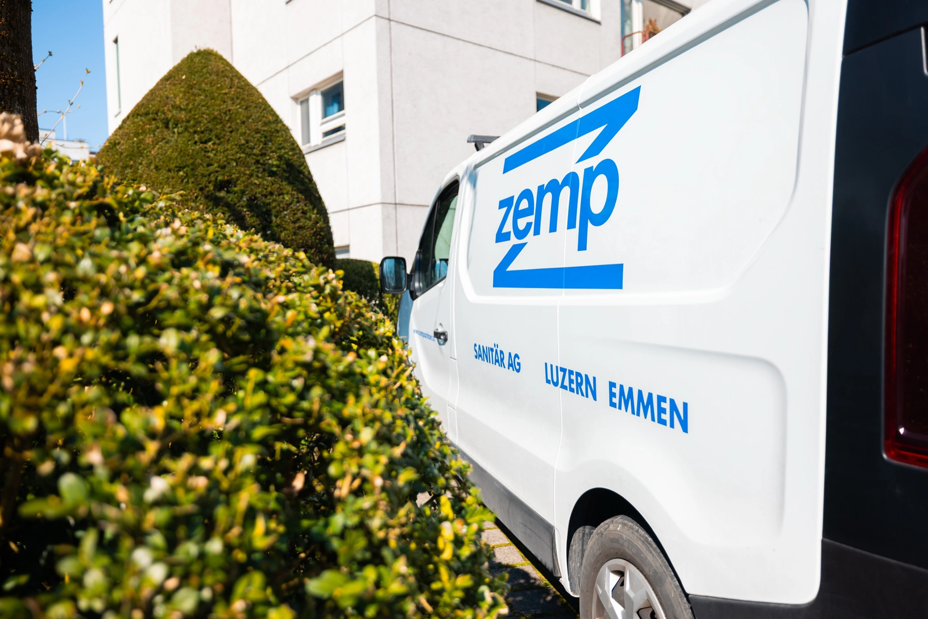 Servicebus der Zemp Sanitär AG mit Firmenlogo vor grüner Hecke und Schweizer Wohnhaus